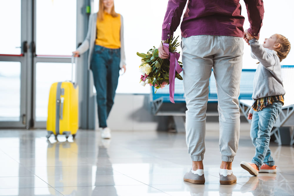 Mann mit Kind begrüßt Frau / Ehegattin am Flughafen mit Blumenstrauß